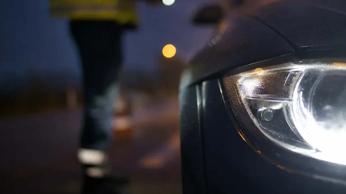Zbliżenie na reflektor samochodu i sylwetkę funkcjonariusza w odblaskowej kamizelce o zmierzchu.
