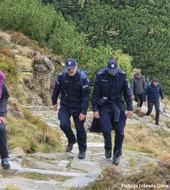 Funkcjonariusze policji i inne osoby wspinają się kamienistą ścieżką w górach porośniętych kosodrzewiną.