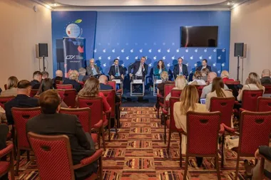 Uczestnicy konferencji lub panelu dyskusyjnego siedzą w sali konferencyjnej przed sceną, na której zasiada panelistów; w tle niebieska ścianka z logo i napisami