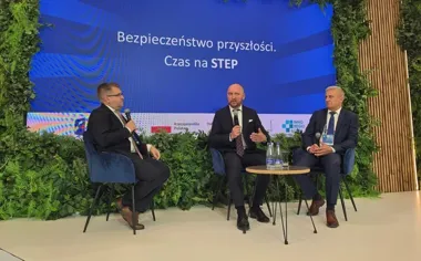 Trzej mężczyźni w garniturach siedzą na niebieskich fotelach podczas panelu dyskusyjnego na tle ekranu z napisem „Bezpieczeństwo przyszłości”.