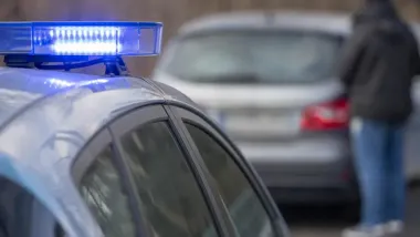Fragment policyjnego radiowozu z niebieskim światłem na dachu, w tle rozmyty samochód osobowy.
