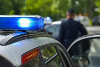 Policyjny radiowóz z włączonym niebieskim światłem, w tle stoi funkcjonariusz przy otwartych drzwiach samochodu.