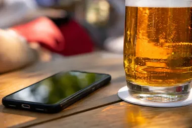 Smartfon i szklanka piwa stoją na drewnianym stole w ogródku kawiarnianym.
