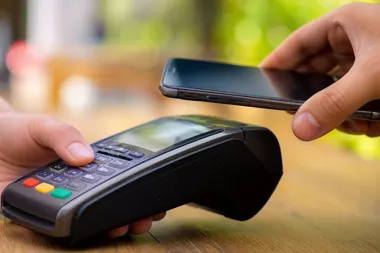 Dwie dłonie przy drewnianym blacie: jedna trzyma terminal płatniczy, druga zbliża smartfon do czytnika NFC.