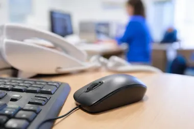 Mysz komputerowa i klawiatura na biurku w biurze, w tle rozmazana osoba pracująca przy komputerze.