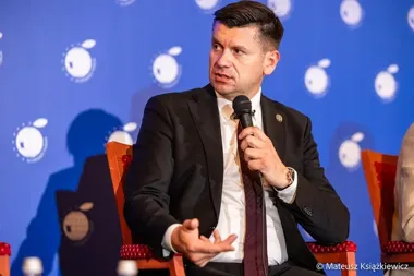 Mężczyzna w garniturze przemawia do mikrofonu na tle niebieskiej ścianki z logotypami.