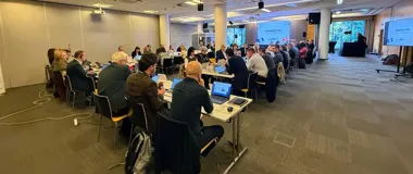 Uczestnicy konferencji lub spotkania biznesowego siedzą przy stołach z laptopami w nowoczesnej sali konferencyjnej.